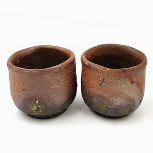 以下が含まれることがあります： 素朴な手作りのセラミックカップ2つ。カップは温かみのある茶色で、表面はざらざらしており、形は少し不規則です。カップの内側は濃い茶色で、底は緑がかった色合いです。