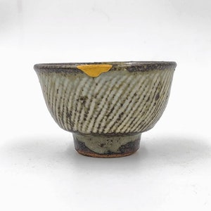 May include: A small, grey ceramic bowl with a textured, striped pattern. The bowl has a brown rim and a small foot.