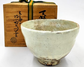 Puede incluir: Una taza de té de cerámica blanca con una superficie rugosa y texturizada y una base marrón. La taza está en una caja de madera con caracteres japoneses en el lateral.