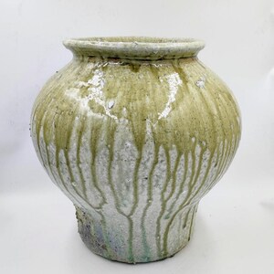 Può includere: Un vaso in ceramica rotondo con un'apertura ampia e una base testurizzata color bianco sporco. La parte superiore presenta uno smalto lucido verde oliva che cola lungo i lati in striature verticali, creando un aspetto stratificato e variegato.