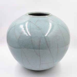 Puede incluir: Un jarrón redondo de cerámica azul pálido con acabado craquelado. El jarrón tiene un cuerpo ancho y esférico y una abertura estrecha en la parte superior. La superficie presenta una red de finas líneas de color marrón rojizo, creando una textura única. Es un objeto decorativo.