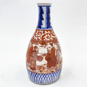 May include: A small, white porcelain vase with a blue and red design. The vase has a narrow neck and a wide, rounded body. The design features a scene of three figures in a landscape, with trees and flowers. The vase is decorated with blue and red lines and swirls.