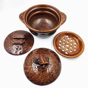 May include: A brown ceramic cooking pot with a textured exterior, featuring a matching lid with a handle, a separate circular insert with multiple drainage holes, and a smooth interior basin with two side handles for easy lifting and handling.