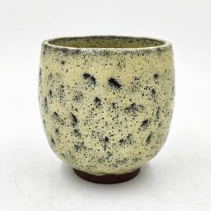 May include: A small, yellow ceramic cup with a speckled black glaze. The cup has a rounded shape and a slightly flared rim.
