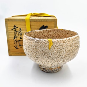 May include: A textured ceramic bowl with a beige and brown speckled pattern and a gold accent. The bowl is next to a wooden box with a yellow ribbon and Japanese characters.