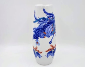 May include: A white ceramic vase with a blue and orange phoenix design. The vase is tall and slender with a rounded bottom.