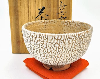 Könnte beinhalten: Eine weiße Teeschale aus Keramik mit einer strukturierten Oberfläche und einem braunen Boden. Die Schale steht auf einem roten Tuch und befindet sich in einer Holzkiste mit japanischen Schriftzeichen.