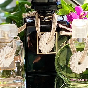 May include: Three glass perfume bottles with decorative white flower-shaped bottle stoppers and brown twine tied around the necks of the bottles. The bottles are sitting on a reflective surface.