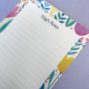 Könnte beinhalten: Ein rechteckiger Notizblock mit weißem, liniertem Papier und einem bunten Blumenrand in Blau-, Gelb-, Rosa- und Grüntönen. Der Text 'Gigi's Notes' ist oben in der Mitte der Seite in Schwarz gedruckt und von stilisierten botanischen Illustrationen umgeben.