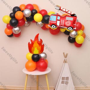 May include: Fire truck, flame, and balloon arch decorations in red, orange, yellow, black, and silver for a birthday party.