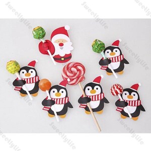 May include: A festive arrangement of Christmas-themed lollipops and cutouts. The image features Santa Claus and penguin cutouts, each holding a lollipop. The penguins wear red and white striped scarves and Santa hats. The lollipops are various colors.