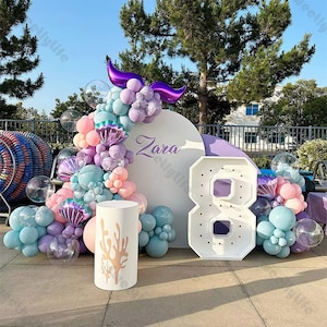 May include: A white round backdrop with the name "Zara" written on it, decorated with a garland of pastel blue, pink, and purple balloons. A large white number 8 with lights is in front of the backdrop. A white cylinder with a coral design is in the foreground.