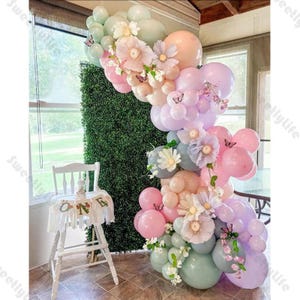 Puede incluir: Un arco de globos en tonos pastel con globos rosas, melocotón, lavanda y verde menta, adornado con flores de papel y mariposas. Una trona blanca con una pancarta que dice "ONE" está en primer plano. El arco está colocado contra un seto verde.