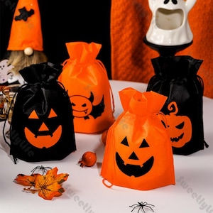 Puede incluir: Una colección de bolsas con cordón de Halloween en negro y naranja. Las bolsas presentan diseños de caras de jack-o'-lantern y diabólicas. También se ven una pequeña araña y una hoja de otoño, junto con un gnomo y una decoración de fantasma.