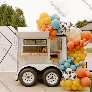 May include: A white food truck with a wooden counter and a window. The truck is decorated with a balloon arch made of yellow, orange, blue, and white balloons. The balloons are arranged in a cascading pattern.