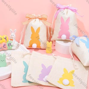 Puede incluir: Bolsas de regalo con temática de Pascua con siluetas de conejos en varios colores, incluyendo naranja, rosa, azul y amarillo. Cada bolsa tiene una cola de pompón blanco y un cierre de cordón. Algunas bolsas tienen lazos decorativos.
