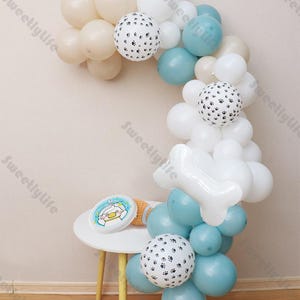 May include: A balloon arch in white, blue, and beige, with paw print and bone-shaped balloons. A small table holds a plate with a cartoon character and a plush toy. The balloons are arranged in an arch shape, suitable for a party or celebration.