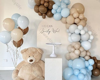 May include: A teddy bear sits in front of a backdrop of blue, brown, and beige balloons. The balloons are arranged in an arch shape. A sign reads "We Can Bearly Wait".