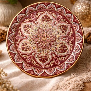 May include: Ceramic decorative plate with a detailed floral design in burgundy, cream, and gold. The plate features an intricate pattern with a gold rim, showcasing a vintage aesthetic. The plate is likely used for display or serving.