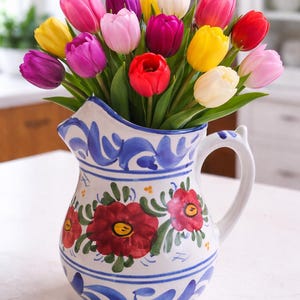 May include: A ceramic jug filled with a vibrant bouquet of tulips. The flowers display a variety of colours, including red, pink, yellow, purple, and white. The jug is white with blue and red floral designs and a handle.