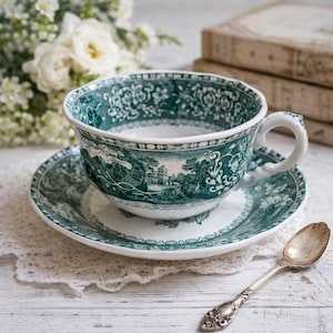 Op de afbeelding: Een vintage kop en schotel set met een groen en wit landschapspatroon. De kop heeft een gebogen oor en staat op een bijpassende schotel. Een zilveren lepel ligt naast de set, wat wijst op een theetijd. De set staat op een wit kanten kleedje.