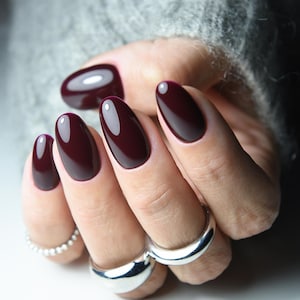 Pode incluir: Uma mão com unhas ovais, bordô escuro e brilhantes. A mão é adornada com anéis de prata, um com design de contas e duas faixas largas e lisas. O fundo é um suéter cinza.