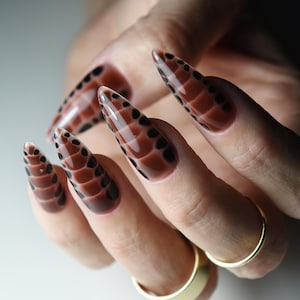 May include: Close-up of a hand with long, pointed nails painted with a brown and black snakeskin pattern.
