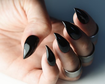 May include: Close-up of a hand with long, black, pointed nails. The nails are painted a glossy black and have a sharp, pointed shape. The hand is wearing several silver rings.