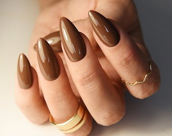May include: Close-up of a hand with five long, almond-shaped nails painted a rich brown color. The nails are glossy and have a smooth finish. The hand is wearing two gold rings.