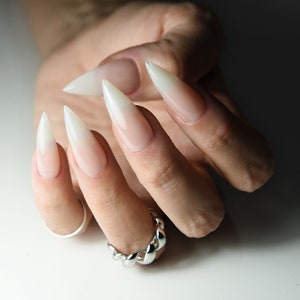 Op de afbeelding: Close-up van een hand met lange, puntige, witte ombre-nagels. De nagels zijn geverfd met een verloop van lichtroze aan de basis naar een witte punt. De hand draagt een zilveren kettingring.