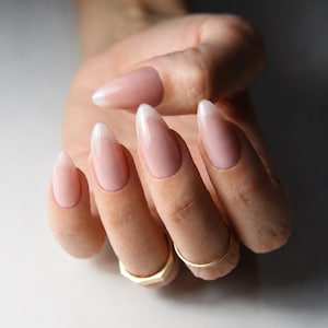May include: Almond-shaped nails featuring a classic French manicure with a sheer, pale pink base and crisp white tips. The nails have a glossy, smooth finish. Two thin, gold-toned metal rings are worn on the middle and ring fingers.