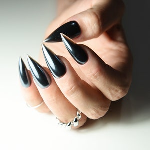 Könnte beinhalten: Nahaufnahme einer Hand mit langen, schwarzen Stiletto-Nägeln mit silberner Glitzer-Spitze. Die Nägel sind mit einem glänzenden schwarzen Lack lackiert.