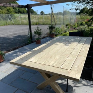 May include: A large wooden table with a natural finish and a cross-shaped base. The table is made of planks of wood and has a rectangular top. The table is sitting on a patio with a pergola overhead.
