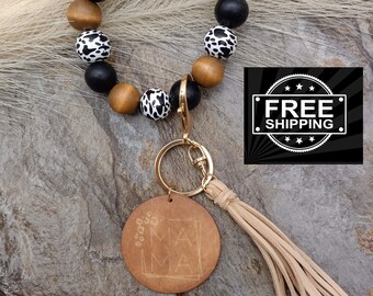 May include: A brown and black beaded keychain with a cow print design. The keychain has a gold metal ring and a brown leather tassel with the word "MAMA" engraved on it.