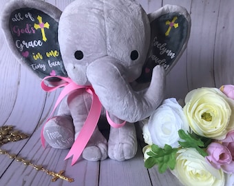 Puede incluir: Un elefante de peluche gris con una cinta rosa alrededor del cuello. Las orejas del elefante tienen texto y un diseño de cruz. Un ramo de flores y un rosario están en el fondo. El texto en la oreja dice "all of God's Grace in one tiny face."