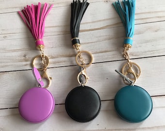 May include: Three leather keychains with round, colorful leather keychains and gold key rings. The keychains have tassels in pink, black, and teal.