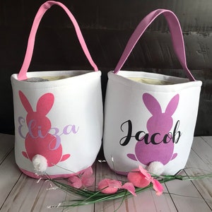 Puede incluir: Dos cestas de Pascua de lona blanca con siluetas de conejos rosa y morado y los nombres "Eliza" y "Jacob" impresos en ellas. Las cestas tienen asas rosas y blancas.