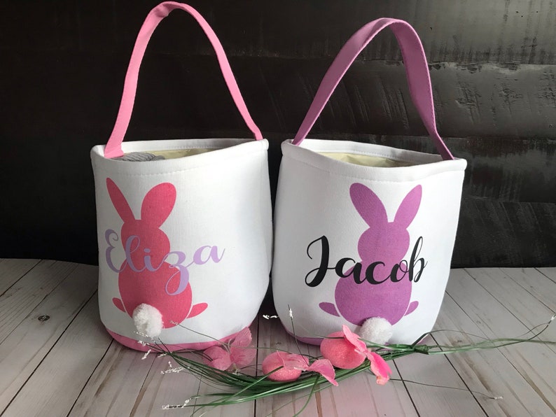 Puede incluir: Dos cestas de Pascua de lona blanca con siluetas de conejos rosa y morado y los nombres "Eliza" y "Jacob" impresos en ellas. Las cestas tienen asas rosas y blancas.