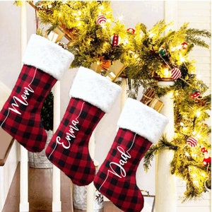 May include: Three red and black buffalo check Christmas stockings with white faux fur cuffs. The stockings are personalised with the names "Mom", "Emma", and "Dad" in white cursive script.