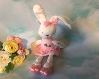 May include: A white plush bunny wearing a pink tutu and pink ballet shoes. The bunny has a pink bow on its head and the name "Mia" embroidered on its chest.