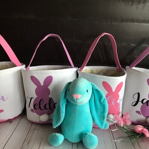Puede incluir: Cuatro cestas de Pascua de lona blanca con siluetas de conejos rosas, morados y azules y nombres personalizados: "Nora", "Ilda", "Liz" y "Jacob". Un conejito de peluche azul claro se sienta frente a las cestas.