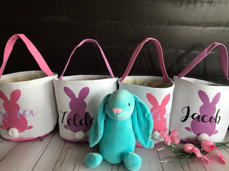 Puede incluir: Cuatro cestas de Pascua de lona blanca con siluetas de conejos rosas, morados y azules y nombres personalizados: "Nora", "Ilda", "Liz" y "Jacob". Un conejito de peluche azul claro se sienta frente a las cestas.