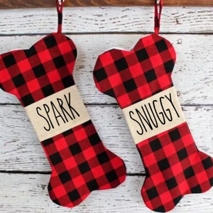 Peut inclure: Deux chaussettes de Noël en forme d'os de chien en plaid buffalo rouge et noir avec les noms "Spark" et "Snuggy" écrits dessus.
