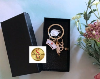 May include: A gold-toned keychain with a white nurse's hat, a syringe, a stethoscope, and a white face mask. The keychain is in a black box with a white background. The box has a gold sticker with the text "Thank you for choosing local. Shelly's Creations. Handmade with love."