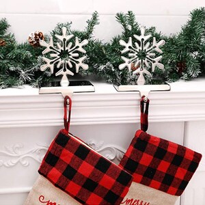 May include: Two silver snowflake stocking holders with a red and black plaid stocking hanging from each. The stockings are hanging from a white mantel.