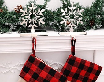 May include: Two silver snowflake stocking holders with a red and black plaid stocking hanging from each. The stockings are hanging from a white mantel.