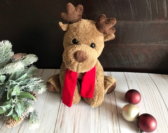 May include: A brown plush reindeer with large, brown eyes and a red scarf. The reindeer is sitting on a white wooden surface with three red and gold ornaments.