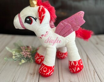 Puede incluir: Unicornio de peluche blanco con alas de purpurina rosa y pezuñas rojas. El unicornio tiene un cuerno dorado y una crin roja. El unicornio está bordado con el nombre "Kaylee" y corazones rojos.