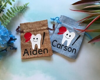 Puede incluir: Dos pequeñas bolsas de arpillera con cierre de cordón. Las bolsas están decoradas con un diente blanco que lleva una gorra de béisbol roja y un bate de béisbol. Las bolsas están personalizadas con los nombres "Aiden" y "Carson".