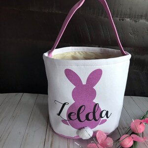 Puede incluir: Una cesta de Pascua blanca con una silueta de conejo morado y el nombre "Zelda" impreso en ella. La cesta tiene un asa rosa y una cola de pomp&oacute;n blanco en el conejo.